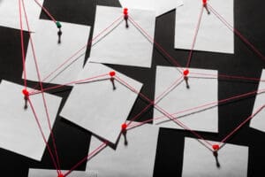 A blackboard with blank white papers pinned to it using red and green push pins. These papers are interconnected by red string, creating a web-like pattern. The arrangement suggests a conceptual network or link between different nodes.