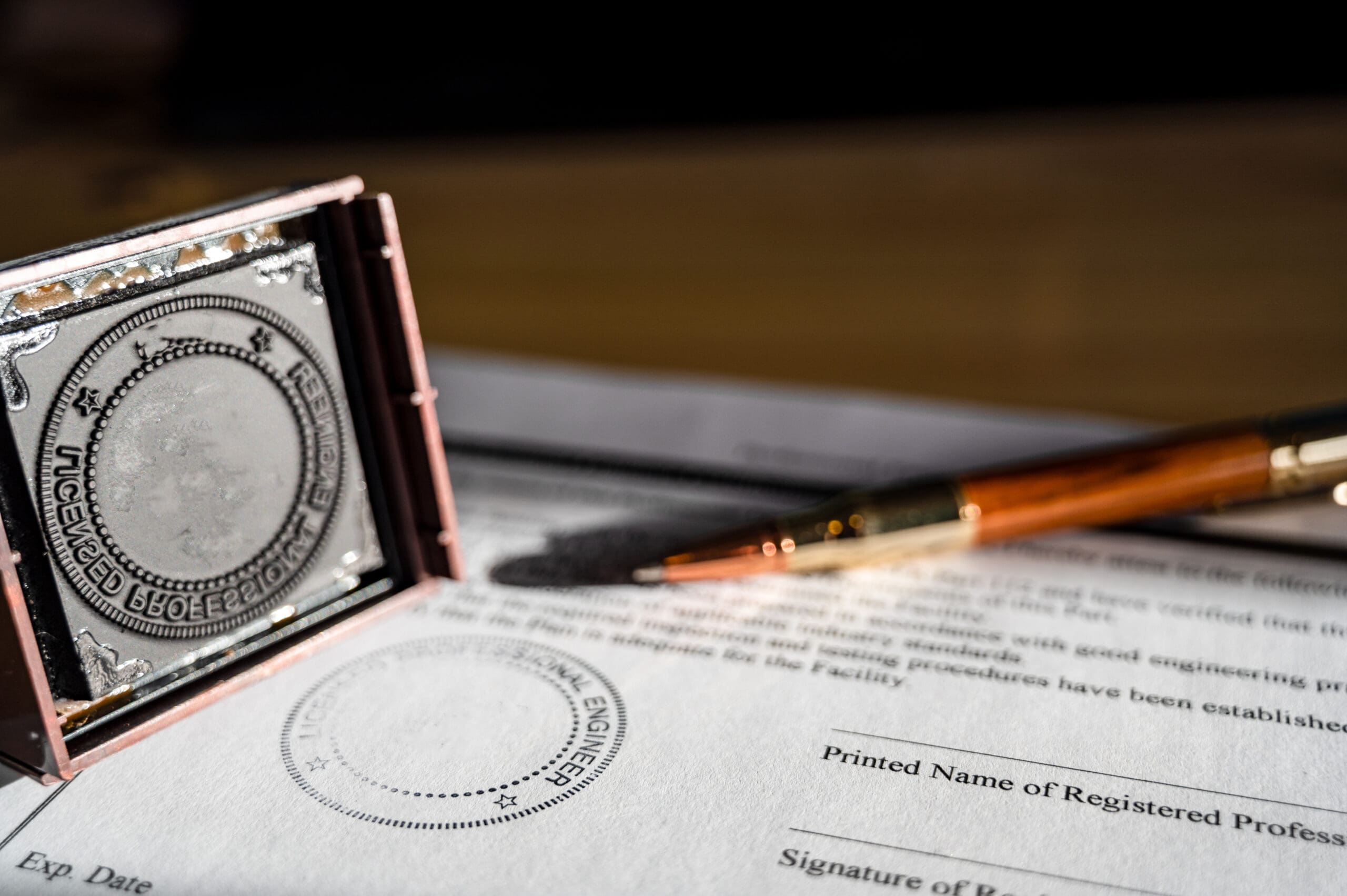 A pen rests on a document next to an engraved professional seal stamp. The document includes spaces for an expiration date, printed name, and signature of a registered professional. The image is well-lit with shadows enhancing the texture of the paper.